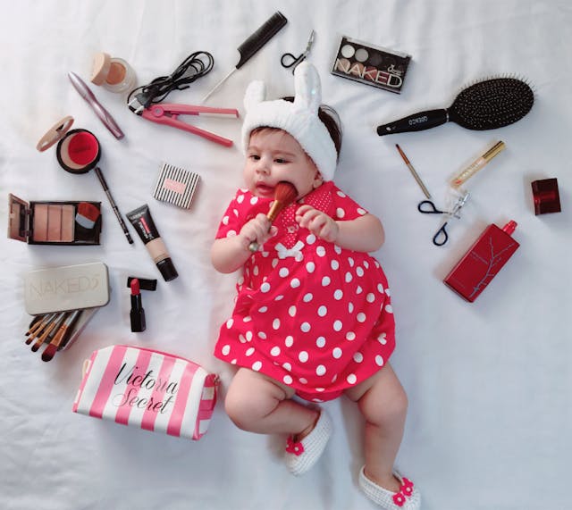 baby surrounded by makeup, hair iron, brushes, and Victoria Secret makeup bag