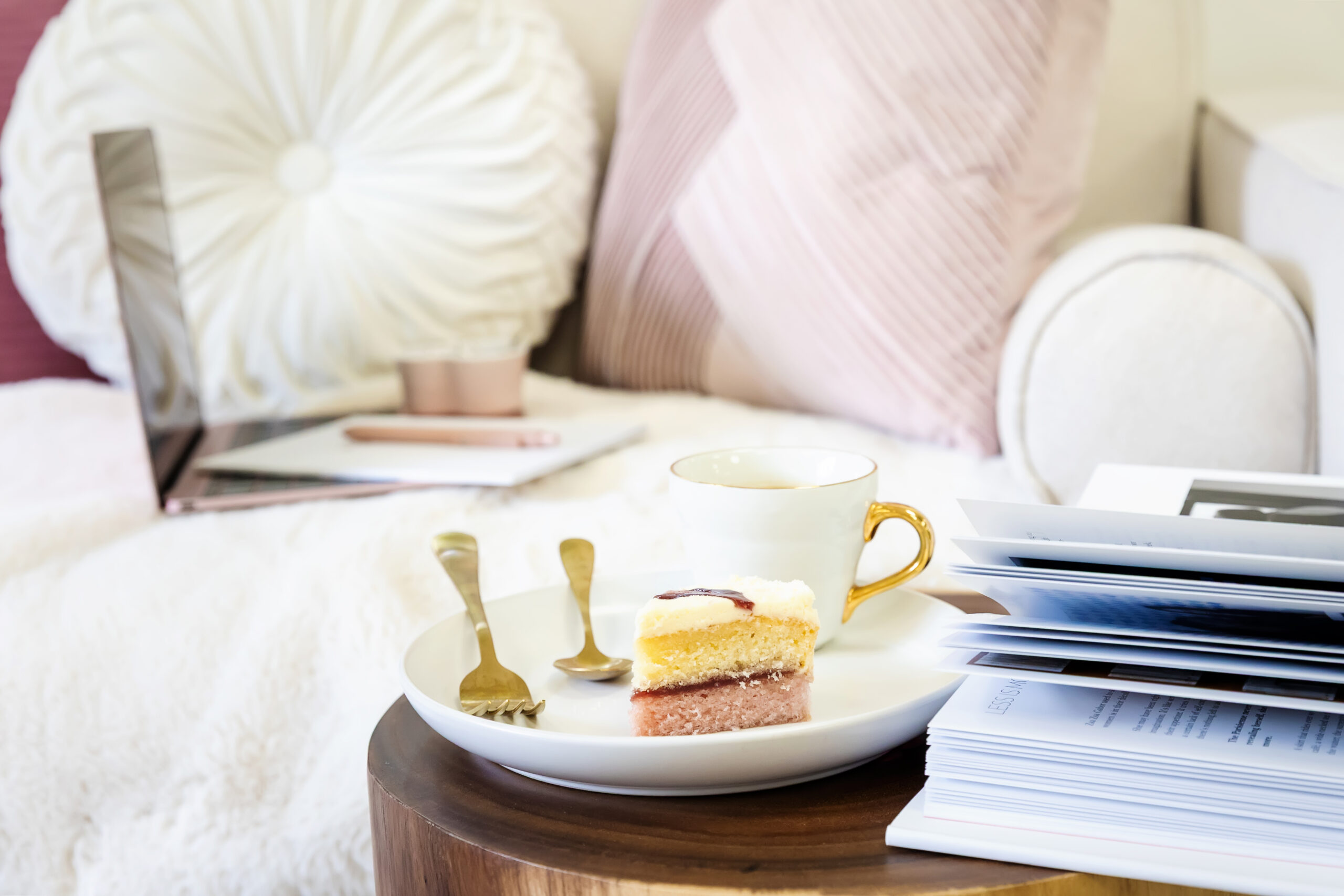 A laptop resting on a cream sofa beside a wooden table with books, a coffee cup, and a slice of cake